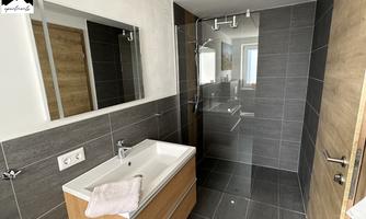 A modern bathroom with dark tiles, a shower, and an elegant wooden vanity. Large windows provide plenty of natural light.