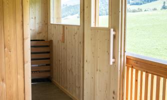 A bright wooden room with open doors, overlooking a green landscape. The walls and floor are made of wood, creating a warm atmosphere.