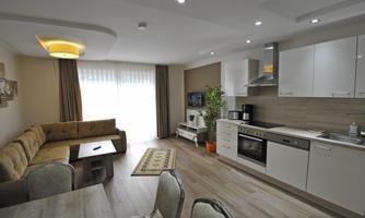 A modern apartment with an open kitchen and living room. Large windows let in plenty of light, and there are cozy furnishings.