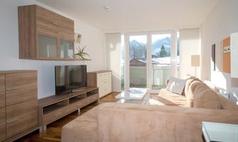A modern living room with a beige couch and a television. Large windows offer a view of the mountains and plenty of natural light.
