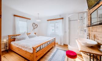 A bright bedroom with a wooden double bed and large windows. In the background, there is a shower and a sink.