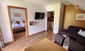 A cozy living and dining room with wooden floors and modern furniture. The room opens up to a bedroom with a double bed.