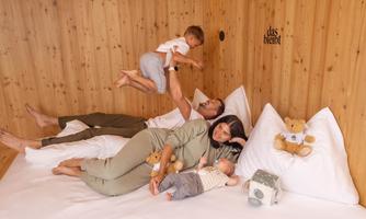 A family is playing on a large bed in a cozy room with wooden paneling. Two children and the parents are laughing and enjoying their time together.