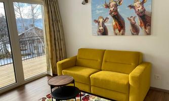 A cozy living room with a yellow sofa and a small table. On the wall hangs a picture of four cows, while large windows provide plenty of light.