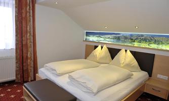 A cozy bedroom with a double bed and comfortable pillows. The walls are subtly designed, and a landscape picture creates a pleasant atmosphere.
