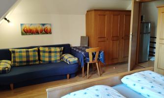 A cozy room with a blue sofa and a small dining table. There is a large wooden wardrobe and two single beds with white bedspreads.