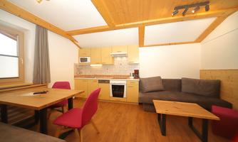 A modern kitchen unit with a dining table and pink chairs. The room is bright and friendly with a wooden ceiling and a cozy sofa.