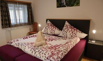 A cozy bedroom with a double bed and decorative cushions. The walls are painted in light tones, and there is a window with curtains.