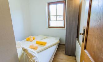 A simple bedroom with a bed, white bedding, and orange towels. The room has a window and a wooden door.