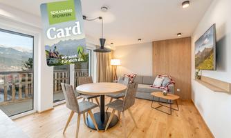 A modern apartment with wooden floors and cozy furnishings. A round table and a couch invite you to relax, while a map of activities in the region is visible.