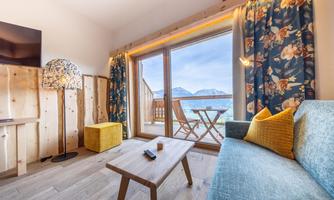 A cozy room with a blue sofa, a coffee table, and large windows that offer a view of the mountains. Decorative curtains and modern furniture give the room an inviting atmosphere.