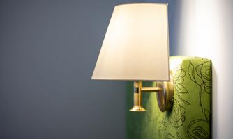 A modern wall lamp with a white shade shines on a green wall. In the background, a simple, neutrally colored wall can be seen.