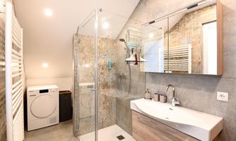 A modern bathroom with a shower and sink. On the left side, there is a washing machine and plenty of daylight from the windows.