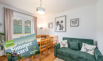 A cozy living space with a dining table and a green sofa. In the foreground, there is a poster with the inscription "Sommercard."