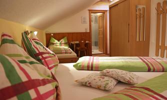 A cozy room with two beds, decorated with colorful cushions and bedding. In the background, there is a small table and chairs as well as a wardrobe.