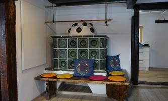 A traditional ceramic stove with colorful cushions on a wooden table in a cozy room. The walls are simply and modernly designed.