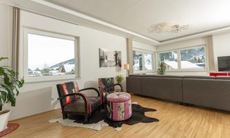 A bright living room with large windows and a view of the mountains. There are colorful chairs and a round table in a modern, minimalist style.