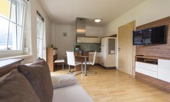 A modern, bright apartment with an open kitchen and a dining area. The room is furnished with a cozy sofa and a television.
