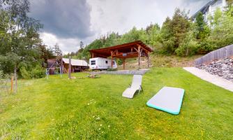 A large garden with a green area, loungers, and a motorhome under a wooden roof. In the background, trees and a path can be seen.