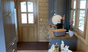 A cozy kitchen in a wooden house with a dining table set for a meal. The wooden paneling and large windows create a bright and warm atmosphere.