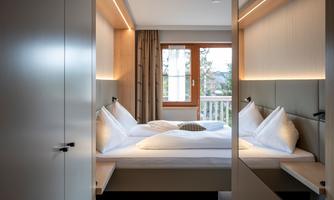 A modern bedroom with two comfortable beds, surrounded by bright walls and warm wood. A large window lets in plenty of daylight and offers a view outside.