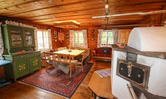 A cozy, rustic room with wooden paneling and a large dining table. The decor includes a traditional stove and a green showcase.
