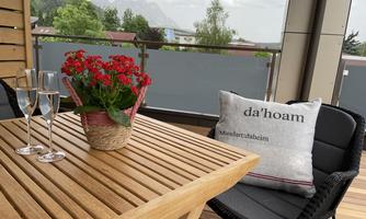 A cozy balcony with a wooden table on which two glasses of sparkling wine are placed. There is also a flower vase with red flowers and a pillow with the print "da'hoam."