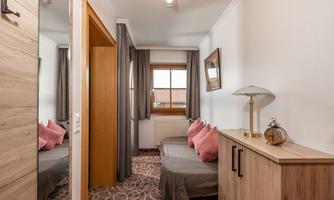 A cozy room with a bed, a wooden wardrobe, and a table. The windows let in a lot of light and the curtains are in a modern style.