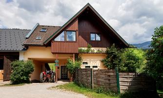 A charming house with wooden elements and large windows. In front of the house is a red tractor, surrounded by green grass and trees.