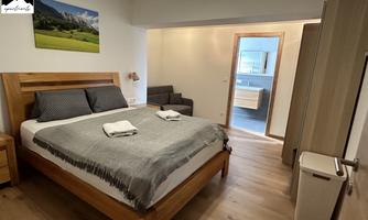 A cozy bedroom with a large bed and soft colors. In the background, there is a modern sideboard and an adjoining bathroom.