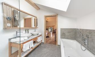 A modern bathroom with a bathtub and two sinks. The space is bright and welcoming, with wood details and gray tiles.