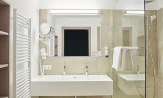 A modern bathroom with a large mirror wall and a vanity. The walls are designed in neutral tones and there is a shower cubicle.
