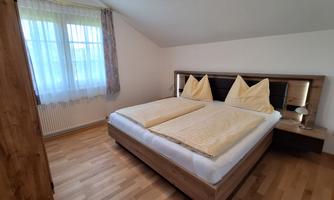 A cozy bedroom with a large double bed and elegant pillows. Bright wooden furniture and daylight coming through the window create an inviting atmosphere.