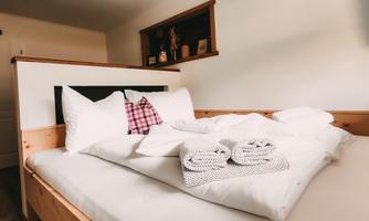 A cozy bed with white bedcovers and pillows. Folded towels lie on the bedspread and a checked pillow is visible.