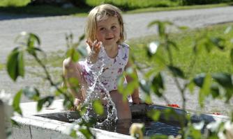A happy child is playing in a water pool and splashing water. In the background, there are green trees and a sunny path.