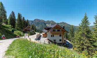 A picturesque mountain landscape with a house and green meadows. In the background, tall mountains and a clear blue sky can be seen.
