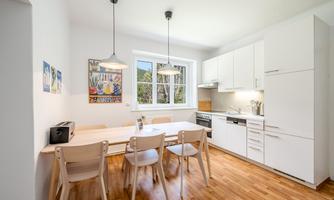 A modern kitchen with a large dining table and bright wood flooring. The walls are white and there are windows overlooking the outdoors.