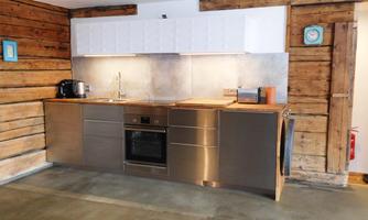 A modern kitchen with stainless steel surfaces and a wood panel. The walls are clad with rustic wood and the furnishings are functional.