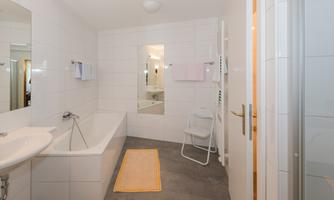 A modern bathroom with a bathtub, sink, and a mirror. There is a rug on the floor and a chair, as well as towels in soft colors.