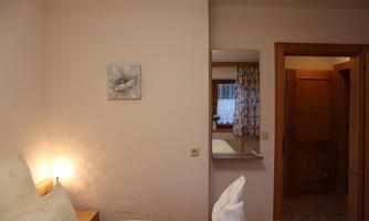 A cozy bedroom with a large bed and soft lighting. In the background, a mirror and access to another room are visible.