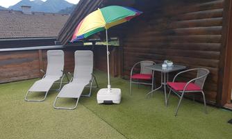 A cozy terrace with two loungers, a colorful sun umbrella, and a small table with chairs. In the background, mountains and a clear sky can be seen.