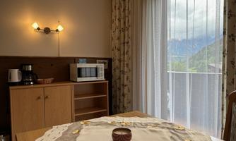 A cozy room with a table and a beautiful tablecloth. In the background, there is a shelf with a microwave and a balcony with a view of mountains visible.