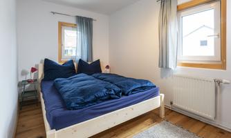 A cozy bedroom with a double bed and blue bedspreads. Daylight streams through the window, and the room has a simple, modern style.
