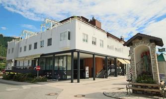 A modern building with a bright facade and large windows. In the foreground, there is a planted area and a bench.