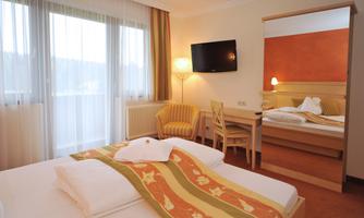 A cozy hotel room with two beds and a small table. Large windows provide plenty of daylight and create a warm, inviting atmosphere.