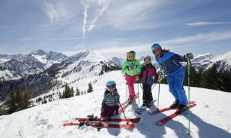A group of skiers stands on a snow-covered mountain with impressive mountains in the background. The sky is clear and blue, and everyone is having fun with winter sports.