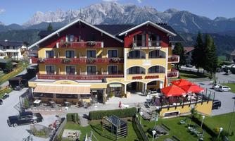 A charming hotel with colorful balconies and an inviting terrace. In the background, majestic mountains and a clear blue sky can be seen.