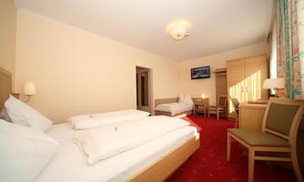 A spacious hotel room with two beds and a small table. The walls are light, and a TV is mounted on the wall.