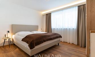 A modern bedroom with a large bed and a cozy wooden floor. Large windows let in plenty of daylight, and the colors are neutral and inviting.