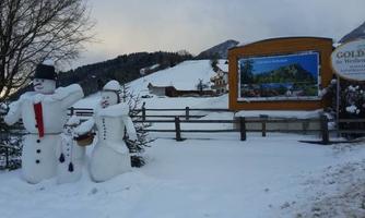 Two snowmen are standing in the snow in front of a sign. In the background, snow-covered hills and trees can be seen.